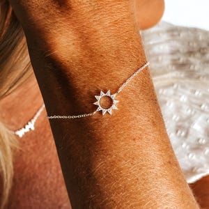 May include: A delicate silver bracelet featuring a sunburst charm with sparkling accents. The bracelet is shown on a person's arm, with the sunburst charm as the focal point. The bracelet's chain is thin and elegant.