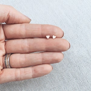May include: A pair of small, gold heart-shaped stud earrings. The earrings are shown on a hand, with the heart-shaped studs placed on the fingertips.