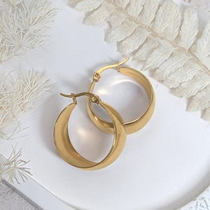 May include: A pair of gold-colored hoop earrings. The earrings are thick and have a smooth, polished surface. They are displayed on a white, textured surface with dried white foliage in the background. The earrings are a classic jewelry item.