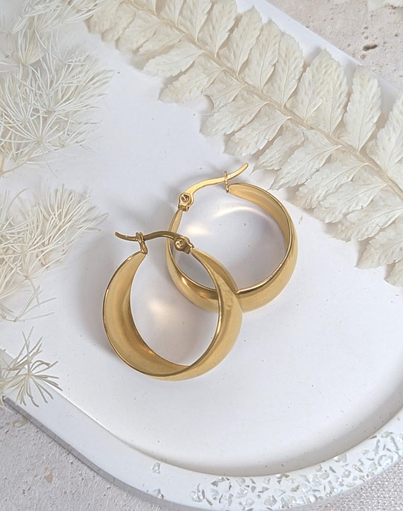 May include: A pair of gold-colored hoop earrings. The earrings are thick and have a smooth, polished surface. They are displayed on a white, textured surface with dried white foliage in the background. The earrings are a classic jewelry item.