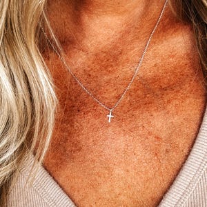 K&ouml;nnte beinhalten: Eine zarte Halskette mit einem silbernen Kreuzanh&auml;nger. Das Kreuz ist klein und h&auml;ngt an einer feinen Kette. Die Halskette wird &uuml;ber einem hellen Oberteil getragen und erzeugt einen schlichten und eleganten Look. Der Schmuck ist ein religi&ouml;ses Symbol.