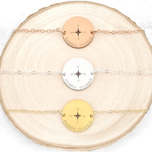May include: Three gold, silver, and rose gold necklaces with compass charms. Each charm has a compass rose and coordinates engraved on it. The necklaces are displayed on a wooden surface.