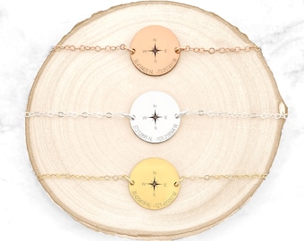 May include: Three gold, silver, and rose gold necklaces with compass charms. Each charm has a compass rose and coordinates engraved on it. The necklaces are displayed on a wooden surface.