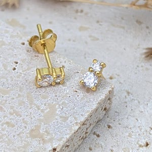 Puede incluir: Un par de pendientes de botón de oro con pequeñas piedras de circonita cúbica transparentes. Los pendientes tienen un diseño sencillo y son perfectos para el uso diario.