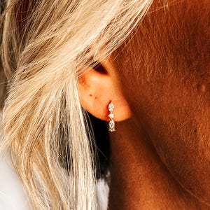 May include: Gold hoop earrings with small, clear gemstones. The earrings are worn in a person's ear.