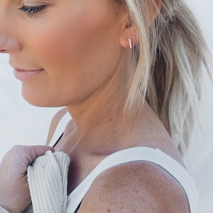 Pu&ograve; includere: Primo piano del viso di una donna con i capelli biondi tirati indietro. Indossa una canottiera bianca e un cardigan marrone chiaro. Indossa piccoli orecchini a cerchio d'argento.