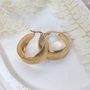 May include: A pair of gold-colored hoop earrings. The earrings are thick and rounded, with a smooth, polished surface. They are displayed on a white tray with decorative elements in the background.