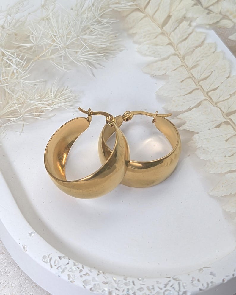 May include: A pair of gold-colored hoop earrings. The earrings are thick and rounded, with a smooth, polished surface. They are displayed on a white tray with decorative elements in the background.