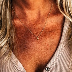 K&ouml;nnte beinhalten: Eine zarte, goldfarbene Halskette mit einem kleinen Kreuzanh&auml;nger. Die Halskette wird um den Hals getragen, wobei das Kreuz in der N&auml;he des Schl&uuml;sselbeins liegt. Der Hintergrund zeigt eine Person, die ein helles Hemd tr&auml;gt.