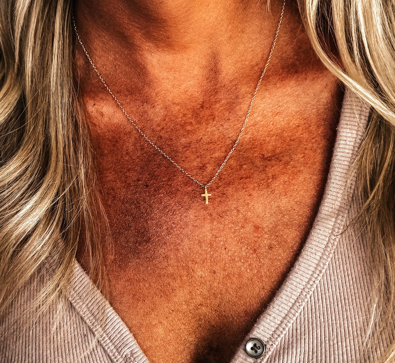 K&ouml;nnte beinhalten: Eine zarte, goldfarbene Halskette mit einem kleinen Kreuzanh&auml;nger. Die Halskette wird um den Hals getragen, wobei das Kreuz in der N&auml;he des Schl&uuml;sselbeins liegt. Der Hintergrund zeigt eine Person, die ein helles Hemd tr&auml;gt.