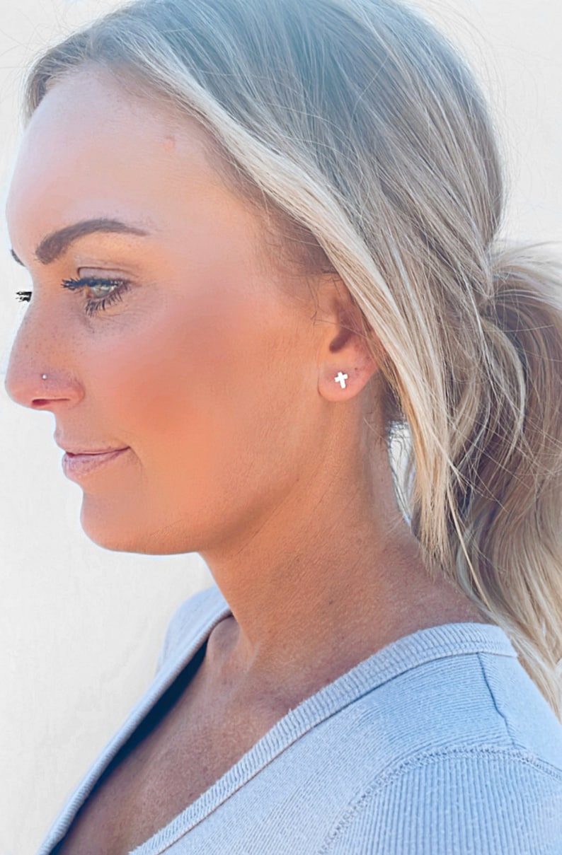 May include: A close-up of a person wearing small silver cross earrings. The person has blonde hair pulled back in a ponytail.