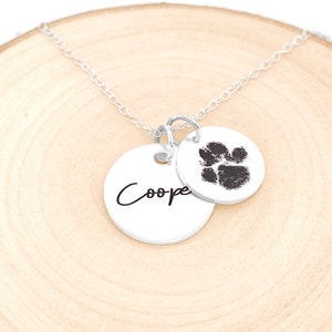 May include: Silver necklace with two charms. One charm is a paw print and the other charm has the name "Cooper" engraved on it.