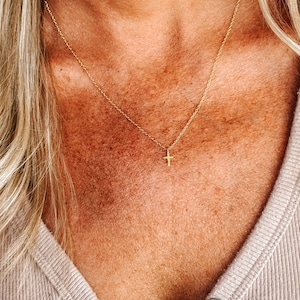 K&ouml;nnte beinhalten: Eine zarte, goldfarbene Halskette mit einem kleinen Kreuzanh&auml;nger. Die Halskette wird getragen, wobei die Kette auf der Haut aufliegt. Das Kreuz ist zentriert, und die Kette ist d&uuml;nn und elegant.