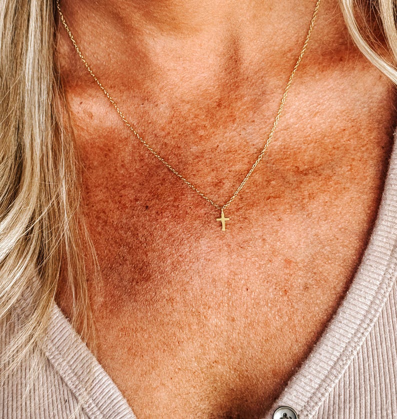 K&ouml;nnte beinhalten: Eine zarte, goldfarbene Halskette mit einem kleinen Kreuzanh&auml;nger. Die Halskette wird getragen, wobei die Kette auf der Haut aufliegt. Das Kreuz ist zentriert, und die Kette ist d&uuml;nn und elegant.