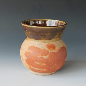 Puede incluir: Un jarrón de cerámica con cuerpo redondeado y borde acampanado. El jarrón está decorado con un esmalte marrón en el borde y un esmalte marrón rojizo en el cuerpo.