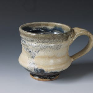 May include: A handmade ceramic mug with a white glaze and blue and black accents. The mug has a rounded body and a handle.
