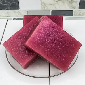 May include: Three bars of pink soap with glitter on a clear glass plate. The soap is a deep pink color with a shimmery finish.