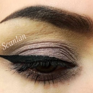 May include: Close-up of a woman's eye with dramatic black eyeliner and eyeshadow. The eyeshadow is a blend of light brown, beige, and purple shades. The word "Scanlan" is written in white text on the eyelid.