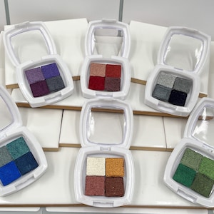 Pode incluir: Seis paletas de sombras brancas, cada uma com quatro tons dispostos em um padrão quadrado. As cores incluem azuis, verdes, roxos e tons terrosos. Cada paleta tem uma tampa transparente com dobradiças.