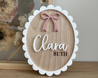 May include: A wooden, scalloped-edge sign with the name "Clara Ruth" in white script and block letters. A pink bow is above the name. The sign is on a wooden surface, with a framed floral arrangement in the background.