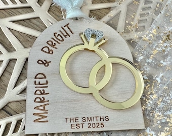 May include: Wooden wedding ornament featuring gold rings and the text "Married & Bright". The ornament includes the names "The Smiths" and the year "EST 2025". A white ribbon is attached for hanging.