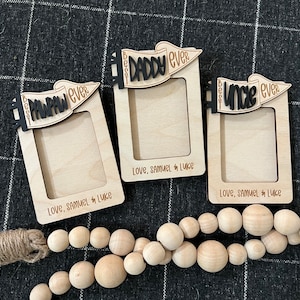 Puede incluir: Tres marcos de fotos de madera con las frases "Best Pawpaw Ever", "Best Daddy Ever" y "Best Uncle Ever" en letras negras y marrones. Cada marco tiene el texto "Love, Samuel & Luke" en la parte inferior. Una guirnalda de cuentas de madera está en primer plano.