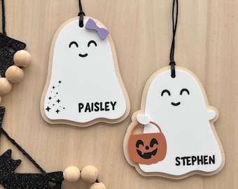May include: Two white ghost-shaped ornaments with black hanging cords. One ghost features a purple bow, black star accents, and the name PAISLEY. The other ghost holds an orange jack-o'-lantern bucket and features the name STEPHEN in black text.