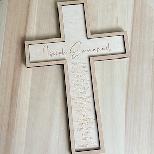 May include: A wooden cross with the words "Isaiah Emmanuel" in a cursive font. The cross features an inscription of Isaiah 41:10. The cross is light brown with a darker brown border and is set against a light wooden background.