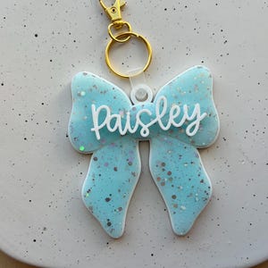 Può includere: Un portachiavi a forma di fiocco azzurro con il nome "Paisley" in corsivo bianco. Il portachiavi ha un moschettone e un anello color oro. Il fiocco è decorato con glitter.