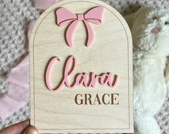 May include: Wooden sign with a light wood grain finish, featuring the name "Clara" in pink script, with "GRACE" engraved below. A pink bow is at the top. The sign is arched at the top and rectangular at the bottom.