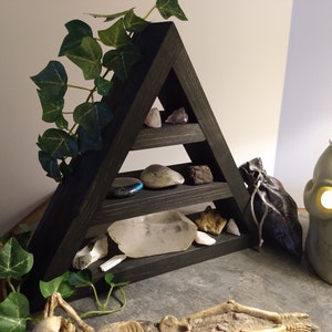 May include: A black, triangular wooden shelf displays various crystals and stones. Green ivy trails down the side. A skull-shaped candle holder and a small bag are also visible, creating a mystical aesthetic.