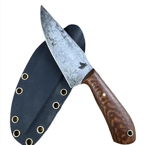 May include: A handmade knife with a dark wood handle and a black leather sheath. The blade is made of steel and has a distressed finish. The knife has a bird logo on the handle.