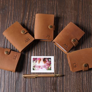 May include: A collection of personalized brown leather wallets with snap closures, each embossed with initials. A small photo and a gold pen are also on the wooden surface.