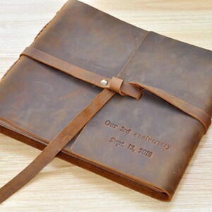 May include: A brown leather photo album with a leather strap and a personalized inscription that reads "Our 3rd anniversary Sept. 12, 2018".