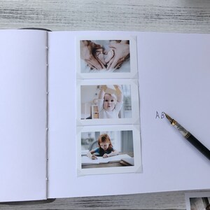 May include: A white photo album with three photos of a baby. The first photo shows the baby's feet in the shape of a heart. The second photo shows the baby sitting in a chair and holding a toy. The third photo shows the baby sitting at a table and drawing. The album has the letter "A" written on the right side.