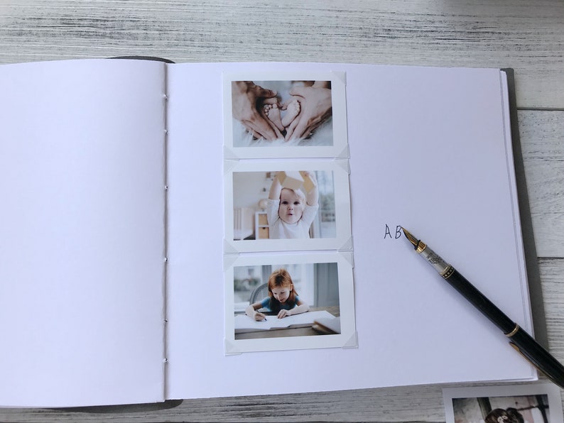May include: A white photo album with three photos of a baby. The first photo shows the baby's feet in the shape of a heart. The second photo shows the baby sitting in a chair and holding a toy. The third photo shows the baby sitting at a table and drawing. The album has the letter "A" written on the right side.