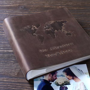 Könnte beinhalten: Ein braunes Fotoalbum aus Leder mit einer geprägten Weltkarte auf dem Einband. Der Text "Our Adventures Tom & Anna" ist unter der Karte geprägt. Ein Foto eines Paares ist im Inneren zu sehen.