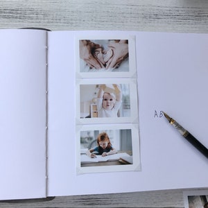 May include: A white photo album with three photos of a baby. The first photo shows the baby's feet in the shape of a heart. The second photo shows the baby sitting in a chair and holding a toy. The third photo shows the baby sitting at a table and drawing. The album has the letters "AB" written on the right side.