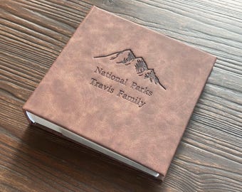 May include: A brown leather photo album with the text "National Parks Travis Family" embossed on the front cover. The album features a mountain range design.