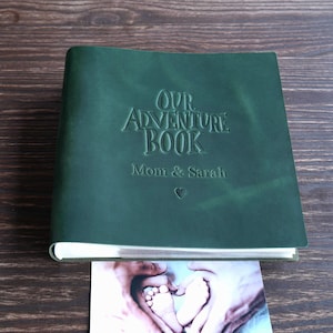 Könnte beinhalten: Ein dunkelgrünes, ledergebundenes Fotoalbum mit dem Titel "Our Adventure Book" mit geprägten Buchstaben. Das Album ist mit "Mom & Sarah" und einem Herzsymbol personalisiert. Ein Foto von Babyfüßen ist sichtbar.