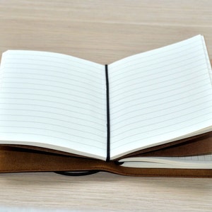 Może przedstawiać: Brązowy notes w sk&oacute;rzanej oprawie z czarną gumką. Notes jest otwarty, ukazując linie na stronach.