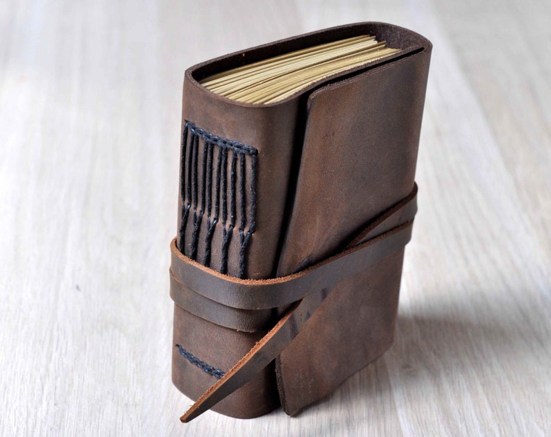 May include: A brown leather-bound journal with a strap closure and black stitching. The journal is closed and lying on a light wood surface.