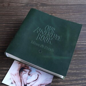 Könnte beinhalten: Ein dunkelgrünes, ledergebundenes Fotoalbum mit dem geprägten Text "OUR ADVENTURE BOOK" und den Namen "Mom & Sarah" darunter. Ein kleines Foto von Babyfüßen ist teilweise sichtbar. Das Album liegt auf einer Holzoberfläche.
