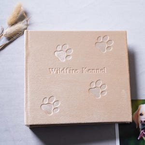 Pode incluir: Um &aacute;lbum de fotos quadrado bege com pegadas em relevo e o texto "Wildfire Kennel". O &aacute;lbum est&aacute; fechado e tem uma apar&ecirc;ncia macia e texturizada. Uma foto de um c&atilde;o &eacute; vis&iacute;vel no canto.