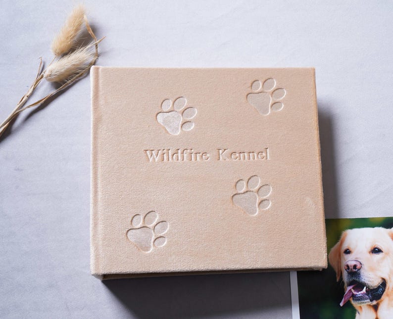 Pode incluir: Um &aacute;lbum de fotos quadrado bege com pegadas em relevo e o texto "Wildfire Kennel". O &aacute;lbum est&aacute; fechado e tem uma apar&ecirc;ncia macia e texturizada. Uma foto de um c&atilde;o &eacute; vis&iacute;vel no canto.