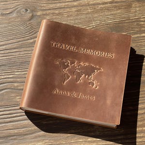 Könnte beinhalten: Ein braunes Leder-Reisetagebuch mit den Worten "TRAVEL MEMORIES" auf dem Einband, einer Weltkarte und den Namen "Anna & James". Das geschlossene Tagebuch liegt auf einer Holzoberfläche.