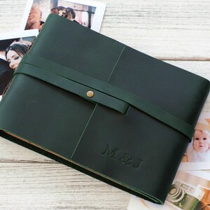 May include: A dark green leather photo album with a brown leather strap and a brass button closure. The album has the initials "M & J" embossed on the front.