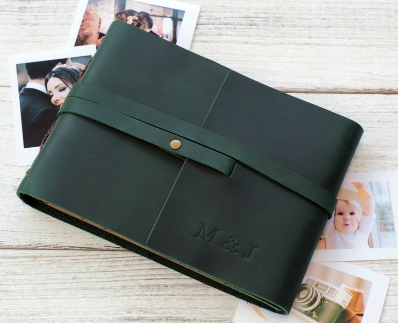 May include: A dark green leather photo album with a brown leather strap and a brass button closure. The album has the initials "M & J" embossed on the front.