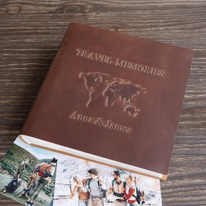 Könnte beinhalten: Ein braunes Leder-Reisetagebuch mit den geprägten Worten "TRAVEL MEMORIES" und "Anna & James" auf dem Einband sowie einer Weltkarte. Das Tagebuch ist geöffnet und zeigt Fotos.