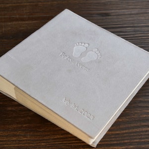 Pode incluir: Um livro de mem&oacute;rias de beb&ecirc; cinza claro com pegadas de beb&ecirc; em relevo e as palavras "Baby Ayers" e "July 1st, 2023". O livro tem uma capa texturizada e uma lombada clara.